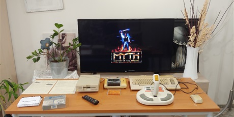 Powiększ grafikę: Na zdjęciu stół, na którym znajduje się komputer Commodore C64 z myszką, joystickiem stacją dyskietek i magnetofonem. Obok leży pudełko z dyskietkami. Przed stołem stoi duży telewizor.