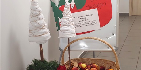 Powiększ grafikę: Na zdjęciu widać drewnianą skrzynkę, na której stoi kosz pełen jabłek. Z tyłu widać świątecznie przystrojoną małą tablicę z informacją o mikołajkach. Z boku stoją ozdoby świąteczne.