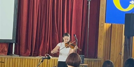 Powiększ grafikę: Dziewczynka na tle sceny, gra na skrzypcach. Zebrana widownia słucha.