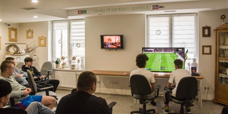 Powiększ grafikę: Dwóch wychowanków siedzących przed telewizorem i grających w grę FIFA20 na konsoli PS4. Z lewej strony na innym telewizorze wyświetlana prezentacja z turnieju fifa z poprzednich lat. 