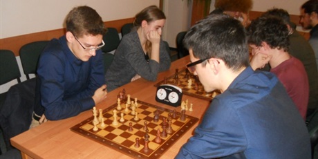 Powiększ grafikę: Zawodnicy siedzą po dwóch stronach stołów i rozgrywając starcie w szachy.