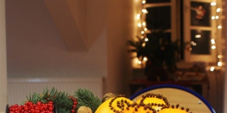 Powiększ grafikę: Na pierwszym planie stół z białym świątecznym obrusem, na którym leżą nabite goździkami pomarańcze i orzechy, przystrojone czerwonym łańcuchem z małych kuleczek, przed nimi garść niebieskich cukierków
