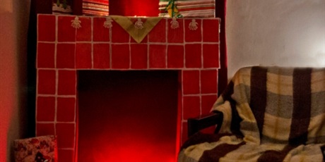 Powiększ grafikę: Czerwony kominek wykonany z kartonu, dookoła niego leżą opakowane prezenty. Na dole rozłożony czerwony łańcuch świetlny. Obok z jego prawej strony fotel przykryty kocem.