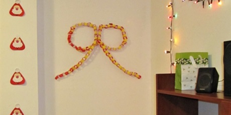 Powiększ grafikę: Kokarda wykonana z łańcucha z papieru, zawieszona na ścianie. Obok po lewej stronie Mikołajki wykonane z papieru, po prawej zaś łańcuch świetlny.