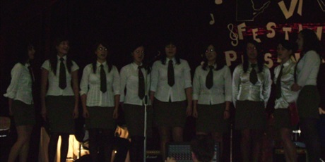 Powiększ grafikę: Dziewięć dziewcząt ubranych w białe bluzki i krawaty występuje na scenie. Za nimi fragment napisu „ VI Festiwal Piosenki”. Przed sceną osoba obsługująca sprzęt muzyczny.