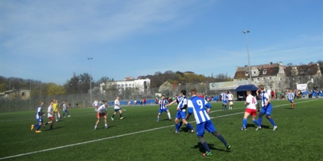 Powiększ grafikę: Toczący się między dwoma drużynami mecz w piłkę nożną. Zawodnicy pierwszej drużyny mają białe stroje, drugiej niebieskie w białe paski.