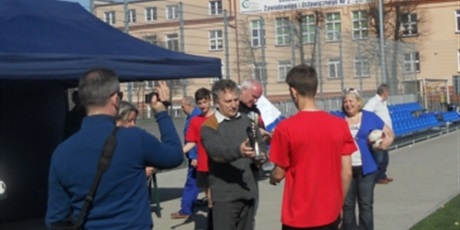 Powiększ grafikę: Dyrektor Bursy Jerzy Serkowski wyciąga rękę w kierunku zmierzającego do Niego, ubranego w czerwony strój zawodnika. W drugiej ręce trzyma puchar.