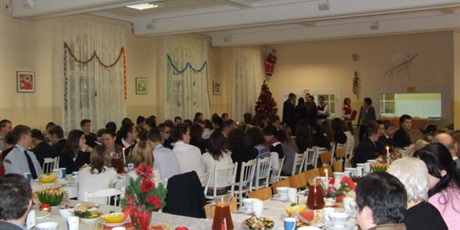 Powiększ grafikę: Wychowankowie i pracownicy wspólnie siedzą za zastawionymi stołami w świątecznie udekorowanej stołówce. Przed nimi występuje grupka młodzieży.