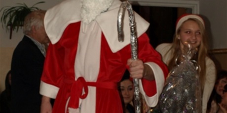 Powiększ grafikę: Święty Mikołaj swoim czerwonym stroju podpiera się o srebrną laskę. Za nim kroczy uśmiechnięta Śnieżynka.