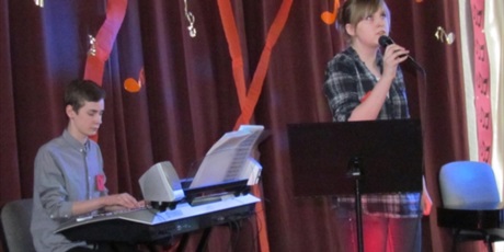 Powiększ grafikę: Na czerwono udekorowanej scenie występuje zespół składający się z dwóch osób. Chłopiec gra na keyboardzie, dziewczynka śpiewa do mikrofonu.