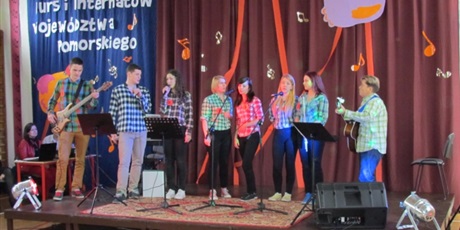 Powiększ grafikę: Na scenie ośmioosobowy zespół, którego członkowie ubrani są w koszule w kratkę. Dwie osoby grają na gitarach, pozostali śpiewają piosenkę. 