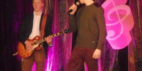 Powiększ grafikę: Na różowo oświetlonej scenie występuje duet. Jeden chłopiec gra na gitarze a drugi śpiewa.