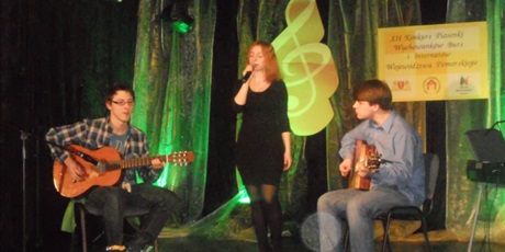 Powiększ grafikę: Na zielono oświetlonej scenie występuję zespół reprezentujący Bursę. Dwóch chłopców gra na gitarach a pośrodku nich  śpiewa wychowanka.