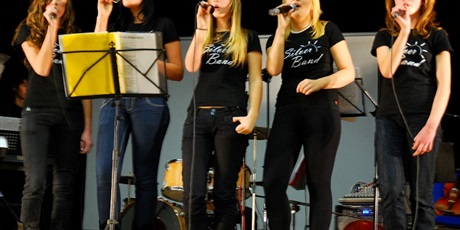 Powiększ grafikę: Piątka dziewcząt występuje na scenie. Ubrane na czarno w rękach trzymają mikrofony do których śpiewają. 