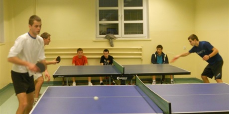 Powiększ grafikę: Zawodnicy rozgrywają mecze przy stołach do pin ponga. Przyglądają im się trzej chłopcy oczekujący na swoją kolej.